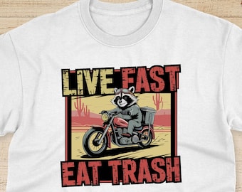 Puede incluir: Una camiseta blanca con un gráfico de un mapache conduciendo una motocicleta en un paisaje desértico. El texto "Live Fast Eat Trash" está impreso en rojo y blanco en la camiseta.