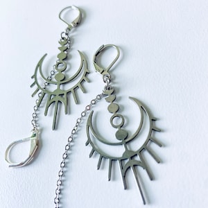 Puede incluir: Pendientes plateados con diseño de luna creciente y rayos de sol, unidos por una cadena. Los pendientes tienen cierre de palanca y una cadena colgante. El diseño incluye pequeños círculos y una línea vertical.