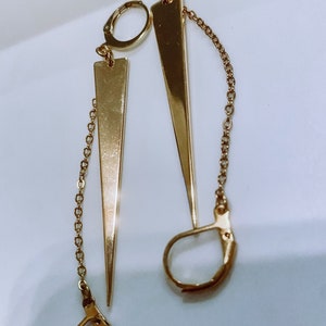 May include: A pair of gold earrings with long, pointed, triangular pendants and dangling chains. The earrings have a simple hoop design.