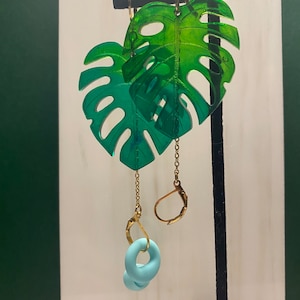 May include: Dangle earrings with translucent green Monstera leaf pendants. The earrings have gold-coloured hooks and chains, with a light blue, donut-shaped charm. The leaves have a gradient effect, going from light to dark green.