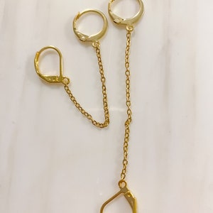 May include: Gold-toned double earrings with a unique design. The earrings feature two hoops connected by delicate chains, with a third hoop and clasp at the bottom. The earrings are displayed on a white marble surface.