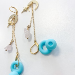 May include: Gold-tone celestial earrings with a crescent moon and star design, dangling chains, and light blue circular and spherical accents. One earring features a pink crystal. The earrings are displayed on a white surface.