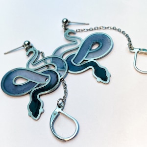 Może przedstawiać: Para srebrnych kolczyków z wzorem niebieskiego i fioletowego węża. Kolczyki są połączone srebrnym łańcuszkiem.