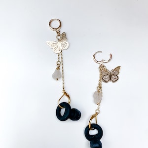 May include: A pair of dangle earrings with gold hoops and accents. Each earring has a gold butterfly charm, a chain with small beads, and a dark blue, circular design. The earrings are set against a white background.