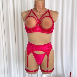 Op de afbeelding: Een levendige fuchsia lingerie set, getoond op een mannequin. De set bevat een bh met open cups, een hoge taille jarretelgordel en een string. De bh heeft decoratieve versieringen. De jarretelgordel heeft verstelbare bandjes en metalen hardware.