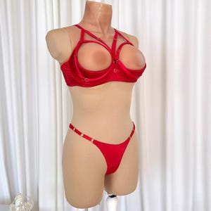 Op de afbeelding: Een rode lingerieset op een mannequin. De set bestaat uit een bh met open cups en een bijpassende string. De bh heeft meerdere bandjes en ringdetails. De string heeft dunne bandjes met ringaccenten. De stof lijkt satijn te zijn.