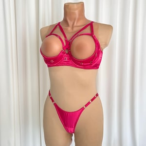 Op de afbeelding: Een felroze lingerieset op een mannequin. De set bestaat uit een beha met open cups en een bijpassende string. De beha heeft decoratieve bandjes en kleine hartvormige accenten. De string heeft dunne bandjes met metalen ringdetails.