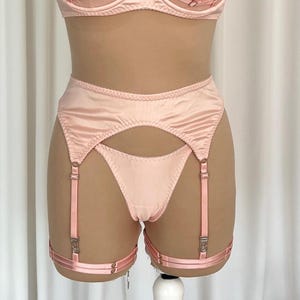 Puede incluir: Conjunto de lencería rosa claro, que incluye un sujetador y una liga de cintura alta con tirantes. La liga tiene un diseño recortado en la parte delantera y correas ajustables. El conjunto está hecho de un material liso, similar al satén.