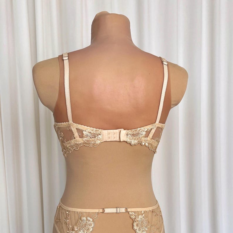 May include: A nude-colored lace bra and panty set with delicate floral embroidery. The bra has adjustable straps and a hook-and-eye closure in the back. The panty has a low-rise cut and a lace trim.