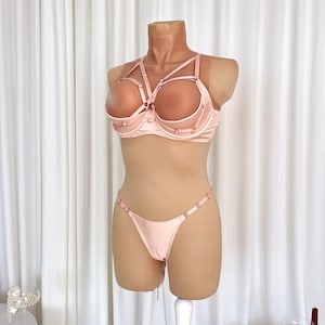 Könnte beinhalten: Hellrosa Dessous-Set auf einer Schaufensterpuppe. Das Set besteht aus einem BH mit einem einzigartigen Trägerdesign und einem passenden String. Der BH hat Bügel und der String verstellbare Träger. Die Dessous sind aus satinartigem Material.