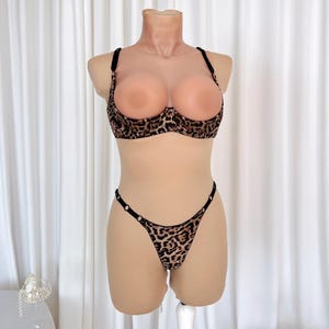 Op de afbeelding: Een set van een beha en string met luipaardprint. De beha heeft zwarte bandjes en beugelondersteuning. De string heeft een dunne tailleband met kleine zwarte versieringen. De set wordt gepresenteerd op een mannequin tegen een witte achtergrond.