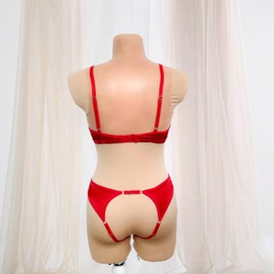 K&ouml;nnte beinhalten: Ein rotes Dessous-Set, bestehend aus einem BH und einem Strumpfhalter. Der BH hat verstellbare Tr&auml;ger und einen Verschluss hinten. Der Strumpfhalter hat verstellbare Tr&auml;ger und einen Verschluss. Das Set wird auf einer Schaufensterpuppe pr&auml;sentiert.