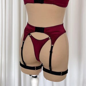 Op de afbeelding: Bordeaux lingerie set met een jarretelgordel, zwarte bandjes en accenten. De set bevat een bijpassende string en een deel van een bh. De jarretelgordel heeft verstelbare bandjes en metalen hardware.