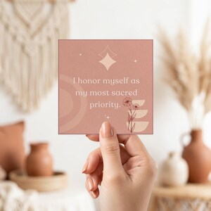 Peut inclure: Une carte carrée rose poudré avec le texte "Je m'honore comme ma priorité la plus sacrée." La carte présente un motif d'étoile, floral et géométrique. La carte est tenue par une main.
