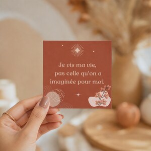 May include: A square, terracotta-colored card with the French text "Je vis ma vie, pas celle qu'on a imaginée pour moi." The card features a white sunburst design and floral accents. The card is held by a person.