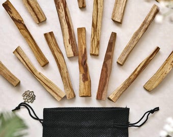 Puede incluir: Una variedad de palos de madera de Palo Santo, de diferentes tamaños y colores, se dispersan sobre una superficie clara. Una bolsa negra con cordón y un dije plateado se encuentran en la parte inferior. Las piezas de madera muestran patrones de vetas naturales, con tonos marrón claro y rojizo.