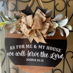 Puede incluir: Un letrero redondo de madera con fondo negro y letras blancas. El letrero dice "AS FOR ME & MY HOUSE we will serve the Lord JOSHUA 24:15". El letrero está decorado con un lazo de arpillera y vegetación.