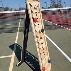 May include: A wooden pickleball scoreboard with black numbers and red and blue scoring pieces. The scoreboard is standing on a tennis court.
