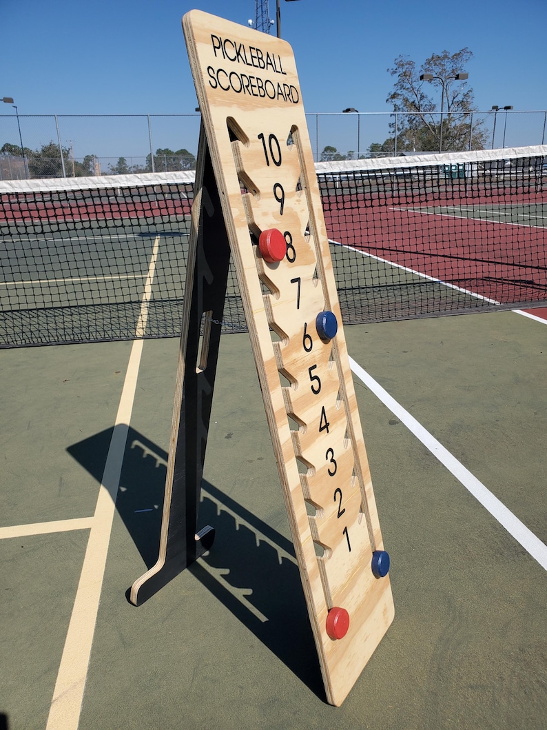 May include: A wooden pickleball scoreboard with black numbers and red and blue scoring pieces. The scoreboard is standing on a tennis court.