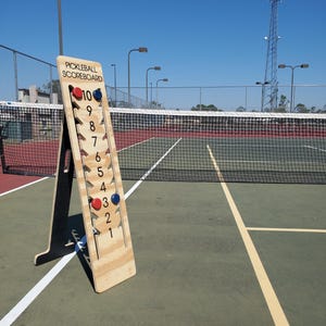 May include: A wooden pickleball scoreboard with red and blue scoring magnets. The scoreboard is on a green court with a tennis net in the background.