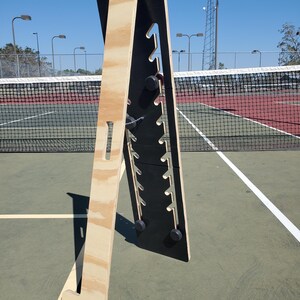 May include: A wooden stand with a black panel featuring multiple slots. The stand is made of plywood and has a T-shaped base. The stand is on a green tennis court.