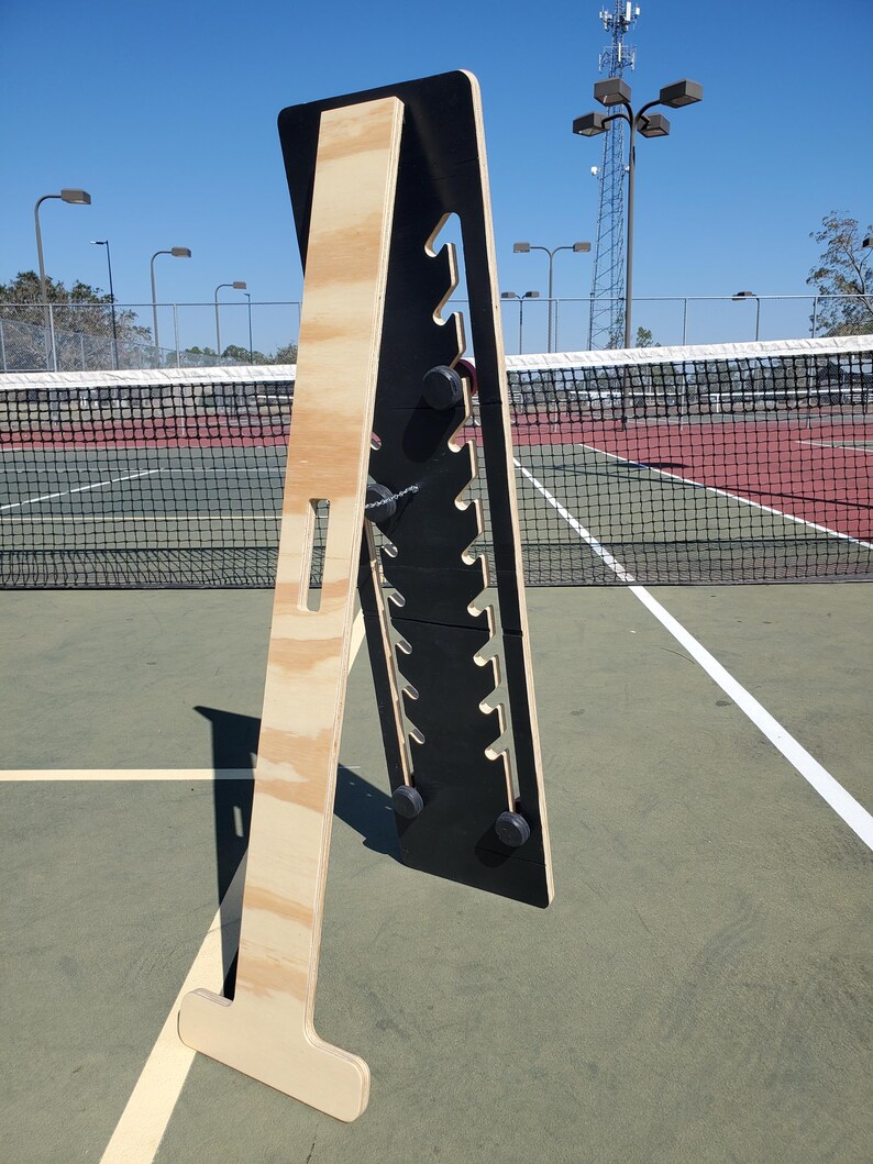 May include: A wooden stand with a black panel featuring multiple slots. The stand is made of plywood and has a T-shaped base. The stand is on a green tennis court.