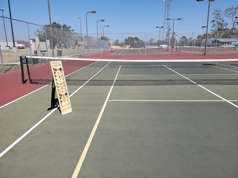 May include: A wooden scorecard for a game of pickleball, with red and blue scoring pieces. The scorecard is on a tennis court with a net in the background.