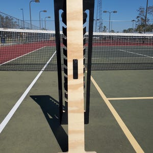 May include: A wooden tennis ball holder with a black frame. The holder has a T-shaped base and a vertical section with slots for holding tennis balls. The holder is standing on a green tennis court.