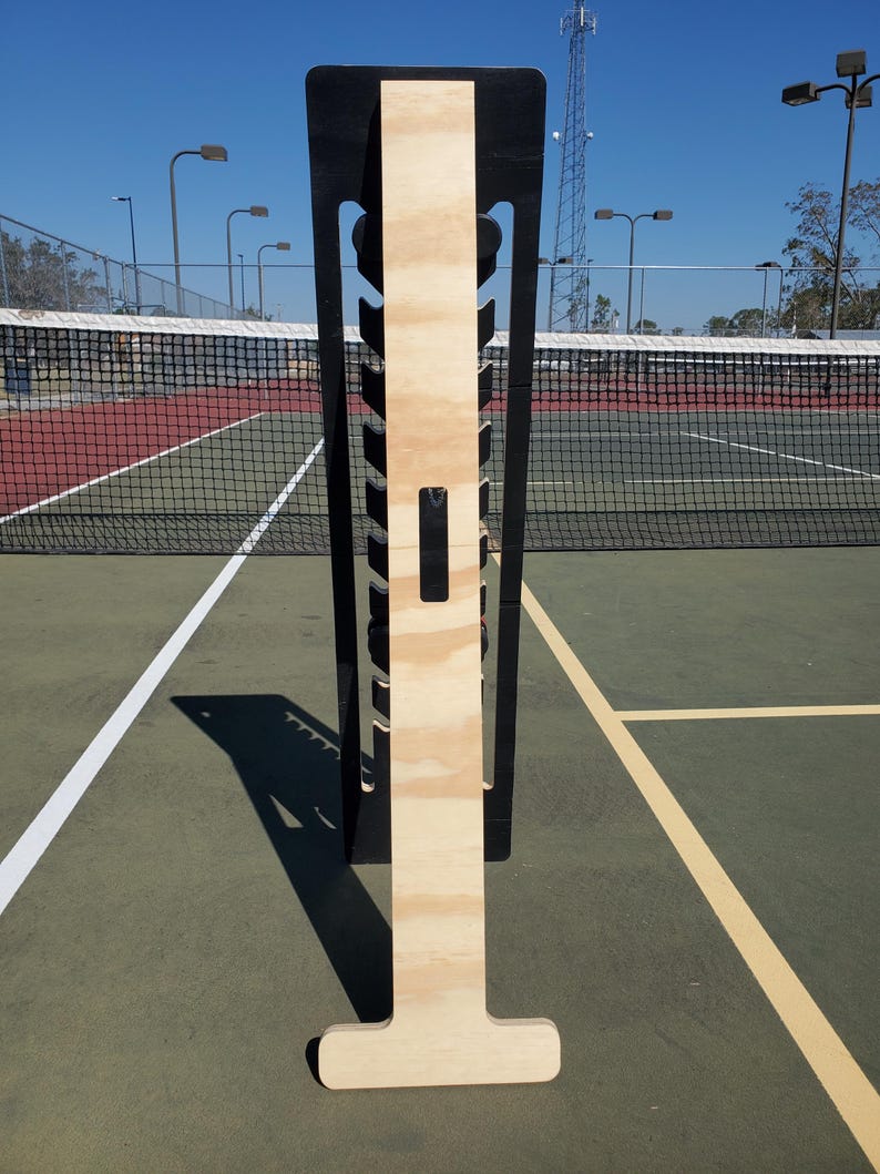 May include: A wooden tennis ball holder with a black frame. The holder has a T-shaped base and a vertical section with slots for holding tennis balls. The holder is standing on a green tennis court.