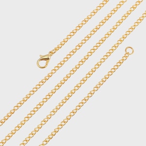 May include: Close-up of several gold-coloured chain necklaces. One chain has a lobster clasp closure, and another has a small ring. The chains are arranged diagonally across the frame, creating a pattern against the light background.