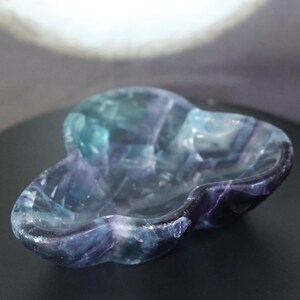May include: A carved fluorite bowl featuring a three-lobed, clover-like shape. The stone displays a natural gradient of deep purple, violet, and translucent teal-green hues with visible internal crystalline structures and smooth, polished edges.