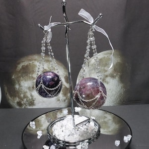 May include: Two spherical, purple and pink gemstone earrings suspended from a silver display stand. Each earring is adorned with a silver chain and a strand of clear beads. White ribbons are tied to the top of the stand.