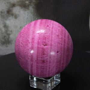 May include: A striking pink sphere with vertical lines, displayed on a clear, square stand. The polished surface of the gemstone reflects light, highlighting its natural patterns. This decorative sphere is a unique home decor item.
