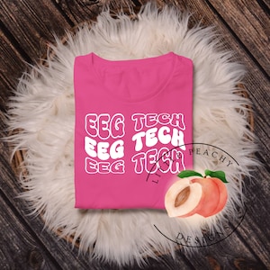 以下が含まれることがあります： 白のレトロなフォントで「EEG TECH」という文字が3回繰り返されたピンクのTシャツ。右下には「Little Peachy Designs」という文字と、半分にカットされた桃が見えます。