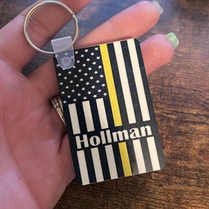 May include: A black and white American flag keychain with a yellow stripe down the middle. The keychain has the name "Hollman" printed on it.