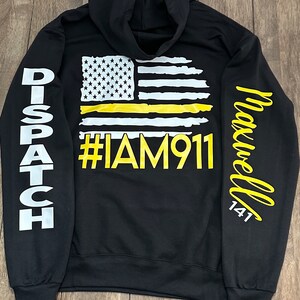May include: Black hooded sweatshirt with a yellow and white American flag with a yellow stripe across the middle. The text "#IAM911" is printed in yellow below the flag. The left sleeve has the word "DISPATCH" printed in white. The right sleeve has the word "Maxwell 141" printed in white.