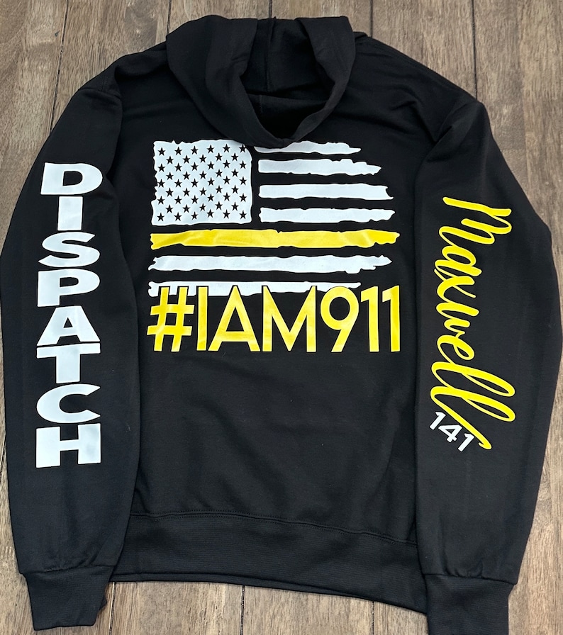 May include: Black hooded sweatshirt with a yellow and white American flag with a yellow stripe across the middle. The text "#IAM911" is printed in yellow below the flag. The left sleeve has the word "DISPATCH" printed in white. The right sleeve has the word "Maxwell 141" printed in white.