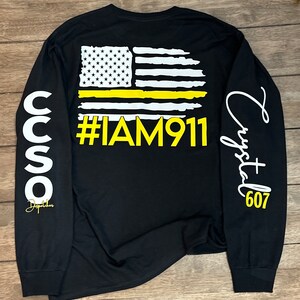 May include: Black long-sleeve shirt with a yellow thin line American flag graphic and the text "#IAM911". The left sleeve has the text "CCSO Dispatcher" and the right sleeve has the text "Crystal 607".