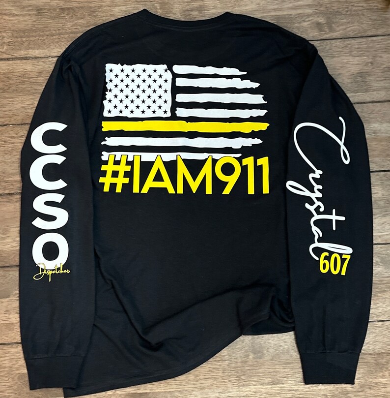 May include: Black long-sleeve shirt with a yellow thin line American flag graphic and the text "#IAM911". The left sleeve has the text "CCSO Dispatcher" and the right sleeve has the text "Crystal 607".