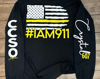 Pode incluir: Camiseta preta de manga comprida com um gráfico de bandeira americana de linha fina amarela e o texto "#IAM911". A manga esquerda tem o texto "CCSO Dispatcher" e a manga direita tem o texto "Crystal 607".