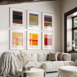 Op de afbeelding: Een woonkamerinterieur met een witte bank en een witte poef. Zes ingelijste abstracte schilderijen met het woord "ROTHKO" erop gedrukt zijn aan de muur gehangen. De schilderijen zijn in tinten rood, geel, blauw en groen.