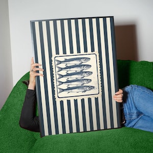 May include: A rectangular artwork featuring a striped pattern in navy and cream. A square image of five fish is centered on the artwork. The artwork is held by a person wearing a black long-sleeve shirt and blue jeans, sitting on a green couch.