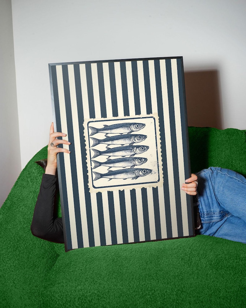 May include: A rectangular artwork featuring a striped pattern in navy and cream. A square image of five fish is centered on the artwork. The artwork is held by a person wearing a black long-sleeve shirt and blue jeans, sitting on a green couch.