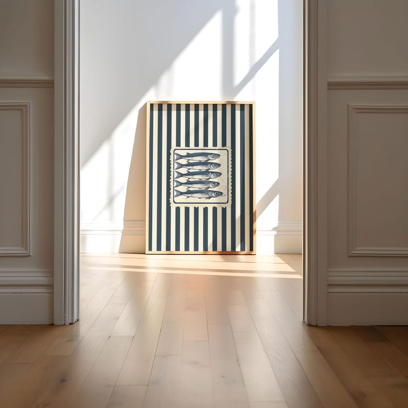 May include: A framed artwork featuring a navy and cream striped background with a central rectangular image of six fish. The artwork is in a gold frame and leans against a white wall, with sunlight illuminating the wooden floor.