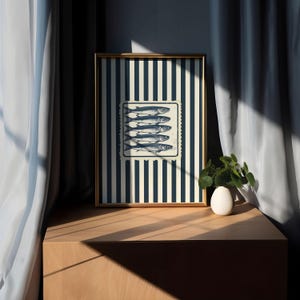 May include: A framed art print featuring a nautical theme. The artwork displays a rectangular image of six fish, set against a striped background of navy blue and cream. The frame is gold-colored, and the print is displayed on a wooden surface.