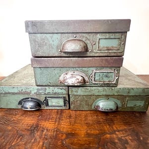 Può includere: Quattro scatole portaoggetti vintage in metallo nei toni del verde e del marrone. Le scatole hanno maniglie e porta etichette. Le scatole sono impilate l'una sull'altra. Le scatole hanno un aspetto invecchiato e usurato.