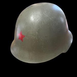 May include: A military-style helmet in a muted olive green colour featuring a prominent red star emblem on the front left side. The surface of the helmet shows visible signs of wear, including various scratches and scuffs across the exterior shell.