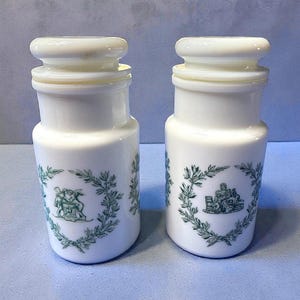 Peut inclure: Deux pots en céramique blancs avec des dessus arrondis et des illustrations vertes. Chaque pot présente un motif de couronne verte autour d'une illustration centrale. Les pots sont sur une surface bleu clair.