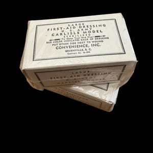 Puede incluir: Dos cajas rectangulares de cartón color hueso de vendaje de primeros auxilios U.S. Army Carlisle Model, envueltas en plástico transparente. El texto dice: Large First-Aid Dressing, U.S. Army, Carlisle Model, Sterilized, Convenience, Inc.