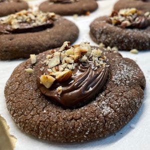 May include: A tray of freshly baked chocolate cookies. Each cookie is round, dark brown, and topped with a swirl of chocolate frosting and chopped nuts. The cookies are arranged on a sheet of parchment paper.